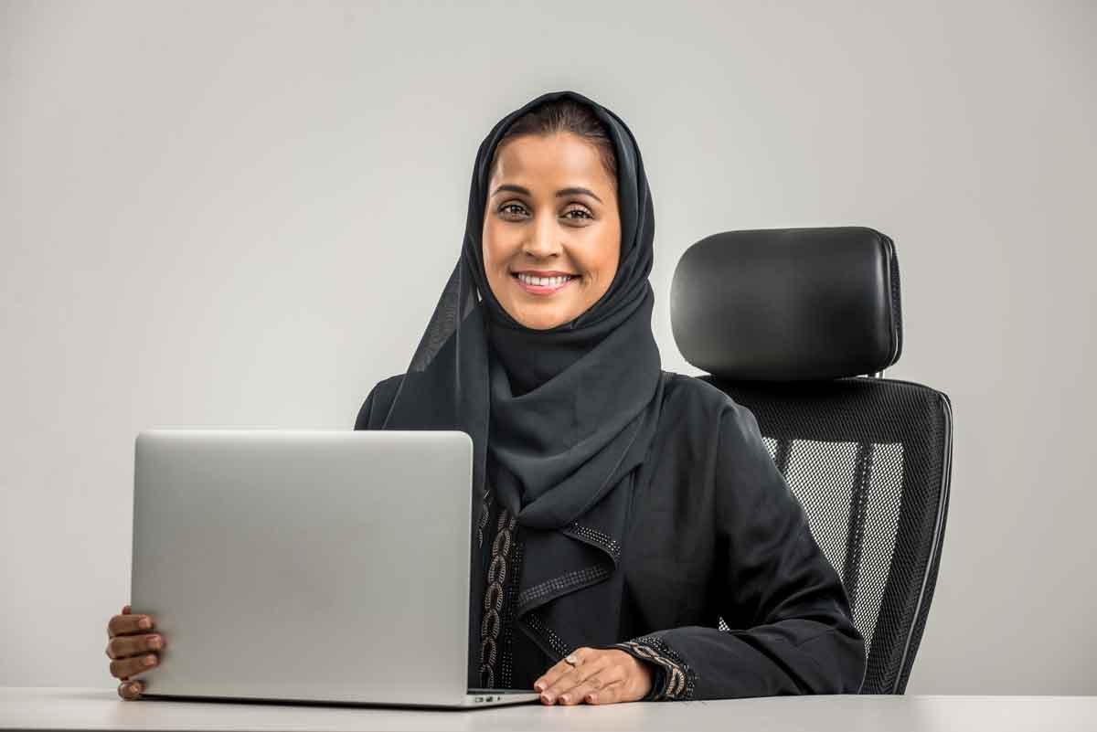 An Arab woman sits on her chair smiling with her laptop at Jawda Plus Institute.